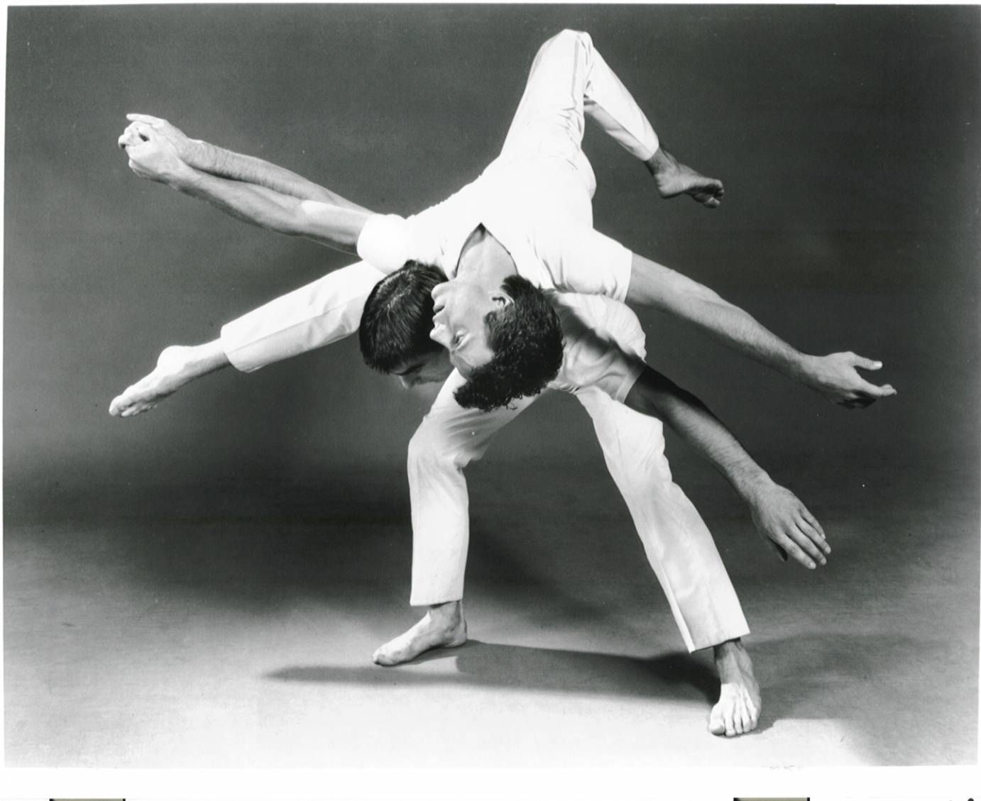 One man rolls over the back of another man, their arms gracefully extended.